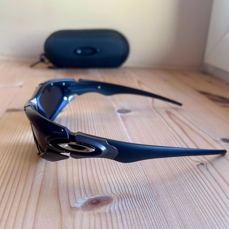 May include: Dark blue sunglasses with a curved frame and dark lenses. The sunglasses are on a wooden surface, with a black case in the background.