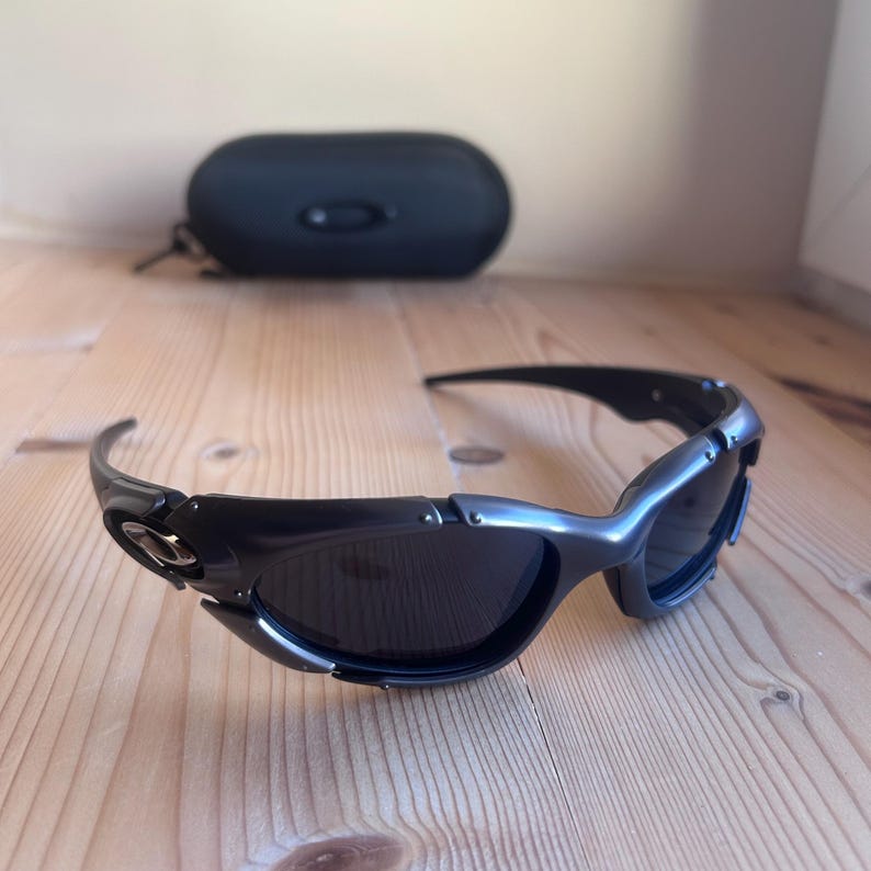 May include: Black sunglasses with a curved frame and dark lenses. The sunglasses are on a wooden surface, with a black case in the background. The frame has silver accents.