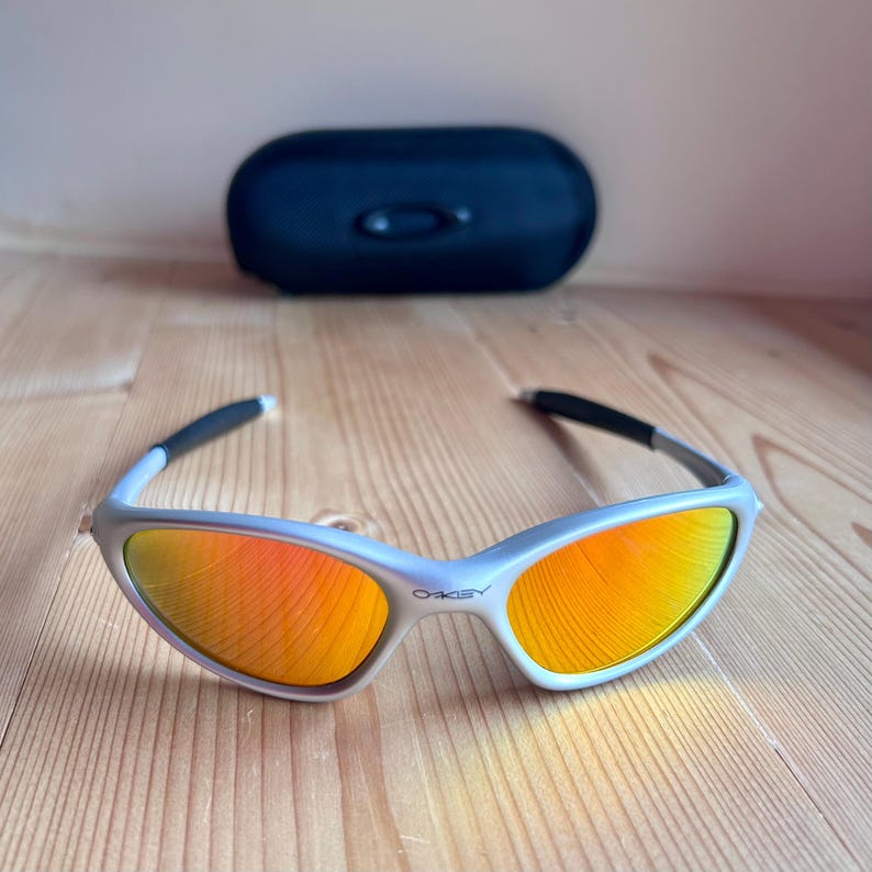 May include: Silver-framed sunglasses with orange lenses and black temples. The sunglasses have the brand name "OAKLEY" printed on the frame. A black, oval-shaped case is in the background. The sunglasses are on a wooden surface.