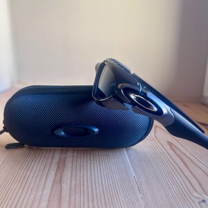 May include: Black sunglasses and a matching black case. The sunglasses have a sleek, modern design with dark lenses and a curved frame. The case is oval-shaped and made of a textured, dark blue material. The sunglasses and case are on a wooden surface.
