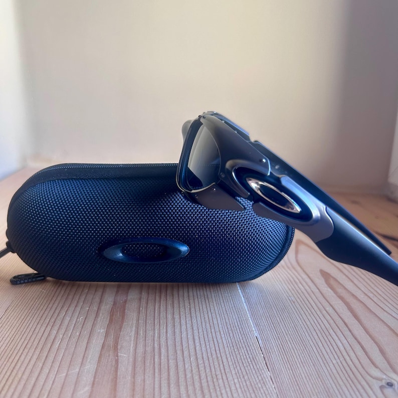 May include: Black sunglasses and a matching black case. The sunglasses have a sleek, modern design with dark lenses and a curved frame. The case is oval-shaped and made of a textured, dark blue material. The sunglasses and case are on a wooden surface.