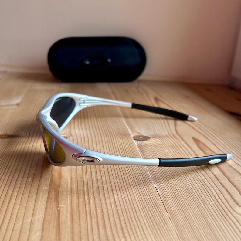 May include: Silver-framed sunglasses with black accents on the temples and a black case in the background. The lenses have a mirrored, multi-colored finish. The sunglasses are resting on a wooden surface.