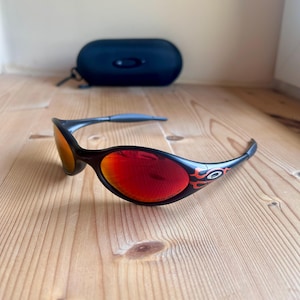 Puede incluir: Gafas de sol negras con lentes rojas y detalles de llamas. Las gafas de sol tienen un diseño curvo y están sobre una superficie de madera. Un estuche negro para gafas de sol está en el fondo.