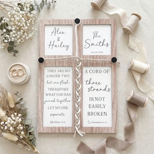 May include: A wooden wedding sign with four framed panels. The top panels read "Alex & Hailey" and "The Smiths June 24, 2026." The bottom panels feature Bible verses about marriage. A braided rope and black accents connect the panels. Gold wedding rings are in a ring box.