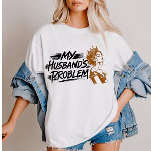 Pode incluir: Camiseta branca com o texto preto "My Husband's Problem" e uma ilustração castanha de uma mulher coroada. A camiseta é de estilo casual, adequada para uso diário.
