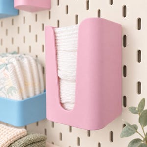 May include: A light pink plastic wall-mounted dispenser containing a stack of white folded cloths. The dispenser features a U-shaped front cutout and is attached to a beige pegboard with vertical slots, positioned next to other storage containers.