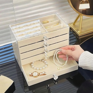 May include: A cream-coloured jewellery box with several drawers and a transparent lid. The box holds various jewellery items, including bracelets, a watch, and rings. The drawers have crystal-like knobs. A person is holding a pearl bracelet.