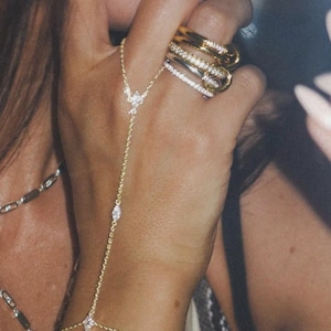 May include: A hand displaying various rings and a gold hand chain. The rings include a gold band with a leopard print pattern, a gold band with clear stones, and a silver band with clear stones. The hand chain is embellished with clear stones.