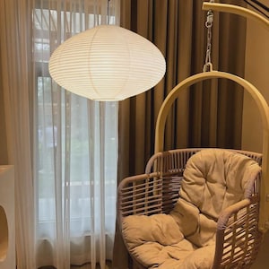May include: A spherical paper lantern hangs above a wicker hanging chair with a tan cushion. The chair is suspended from a curved metal frame. The background features a window with sheer curtains and brown drapes. The lantern is a soft, off-white colour.