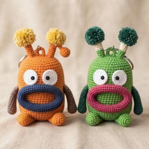 May include: Two crochet monster keychains with metal rings. The left one is orange with a dark blue mouth, brown arms, and yellow pom-poms. The right one is green with a pink mouth, dark green arms, and dark green pom-poms. Both have white eyes.