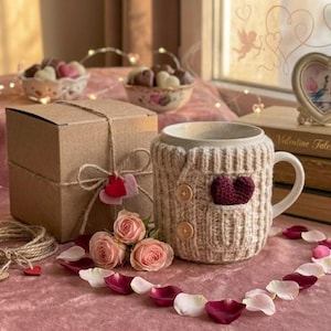 May include: A beige ceramic mug with a knitted sweater sleeve featuring a burgundy heart and wooden buttons. A small gift box tied with twine and a heart-shaped tag sits nearby, along with rose petals and chocolates.