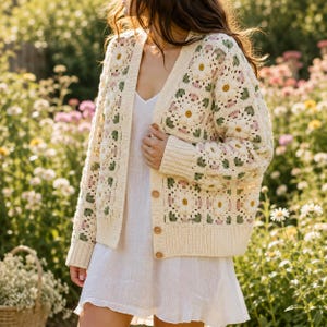 May include: A cream-colored crochet cardigan featuring a granny square pattern with floral motifs in white, yellow, pink, and sage green. The cardigan has a V-neckline, long sleeves, a ribbed hem, and is fastened with four round brown wooden buttons.
