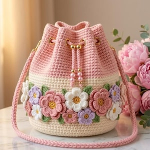 May include: A handmade crochet bucket bag in pink, cream, and white, adorned with colorful crochet flowers. The bag features a drawstring closure, gold-tone hardware, and a pink shoulder strap. A charming and unique accessory.