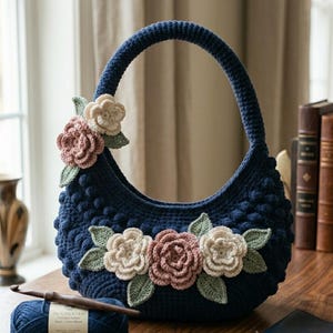 May include: A navy blue crochet handbag with a rounded shape and a sturdy handle. The bag is adorned with crocheted flowers in shades of pink, cream, and green. The bag has a textured surface with small, raised bobbles.