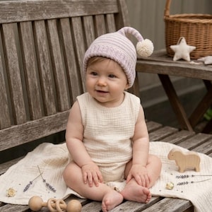 Könnte beinhalten: Ein Baby mit einer hellvioletten Strickmütze mit einem weißen Bommel und einem ärmellosen cremefarbenen Body. Das Baby sitzt auf einer Holzbank mit einer cremefarbenen Decke. Ein Holzschafspielzeug ist ebenfalls sichtbar.