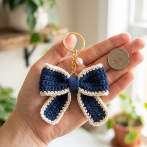 May include: A navy blue and cream crochet bow keychain with a gold-colored ring and a small pearl charm. The bow is held in a hand next to a quarter coin. The bow is a decorative accessory.