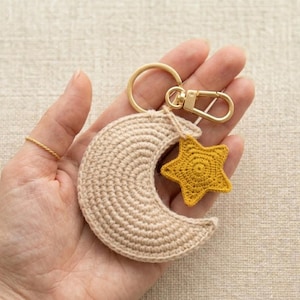 Puede incluir: Un llavero de ganchillo con una luna creciente beige y un dije de estrella amarilla. El llavero tiene un cierre y un anillo dorados. Los artículos están hechos de hilo y probablemente hechos a mano.