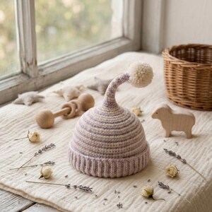 May include: A lavender and cream striped knitted baby hat with a cream pom-pom. The hat is part of a collection of baby items, including a wooden rattle, a wooden sheep, and a small basket, arranged on a white surface near a window.