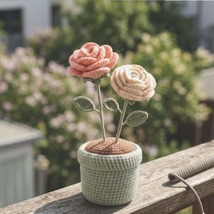 Puede incluir: Una maceta de ganchillo con dos rosas. Una rosa es color melocotón y la otra beige claro. La maceta es verde claro y la base marrón. Las flores tienen tallos y hojas verdes. La maceta está sobre una superficie de madera.