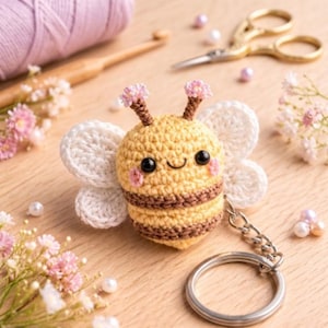 May include: A handmade crochet bee keychain with a yellow body, brown stripes, and white wings. The bee has black eyes, pink cheeks, and small pink flower accents on its antennae. A silver keyring is attached.