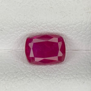 May include: A cushion-cut ruby gemstone with a deep red colour. The gemstone is set against a white textured background. The ruby's facets reflect light, creating a sparkling effect. This gemstone is suitable for jewellery.