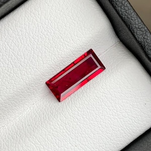 May include: A rectangular, deep red gemstone with a faceted cut, presented in a black jewelry box with a white interior. The gemstone's color is a vibrant, saturated red, and the cut allows light to reflect, creating a sparkling effect.