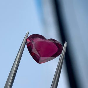 Pode incluir: Uma pedra preciosa vermelha escura facetada em forma de coração é segurada por uma pinça prateada contra um fundo azul suave. As facetas da gema capturam a luz, criando um efeito brilhante. Ideal para fazer joias.