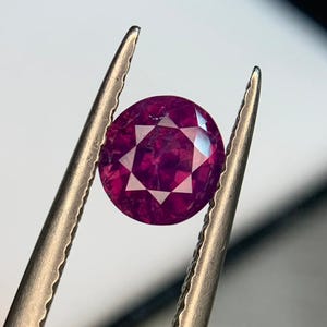 Può includere: Una gemma sfaccettata, rotonda, rosso intenso, è tenuta da una pinzetta d'argento. La gemma ha un motivo a stella di sfaccettature, che riflette la luce. Lo sfondo è di un colore neutro e morbido.