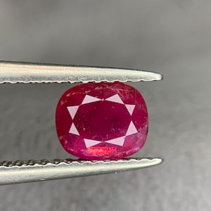 Puede incluir: Una gema de rubí facetada, de forma ovalada, sujeta con unas pinzas. El rubí es de color rojo intenso con un patrón en forma de estrella en el centro. Las pinzas son de color plateado y el fondo es gris neutro.