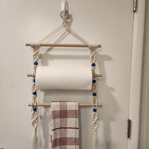 May include: A macrame wall hanging organizer featuring three horizontal wooden dowels suspended by off-white cotton cord. The design includes blue beads, a roll of white paper towels on the middle dowel, and a red and white plaid towel on the bottom.
