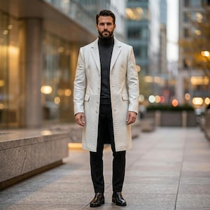 May include: A cream-coloured, textured, long coat with a button closure. The coat is worn with a dark grey turtleneck sweater and black trousers. The outfit is completed with black leather shoes. The background is a city street.