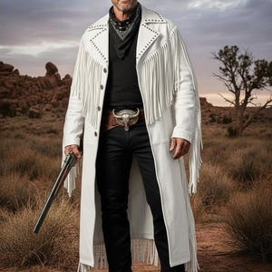Könnte beinhalten: Eine Person trägt einen schwarzen Cowboyhut, ein schwarzes Hemd und eine schwarze Jeans, mit einem weißen Fransenmantel und einer dekorativen Gürtelschnalle. Die Person hält ein Gewehr in einer Wüstenlandschaft.