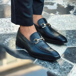 Könnte beinhalten: Ein Paar schwarze Leder-Loafer mit goldfarbener Schnalle. Die Schuhe haben einen strukturierten Vamp und ein klassisches Slip-on-Design. Das Bild zeigt die Schuhe aus einem niedrigen Winkel, der ihre schlanke Silhouette und ihr poliertes Finish hervorhebt.