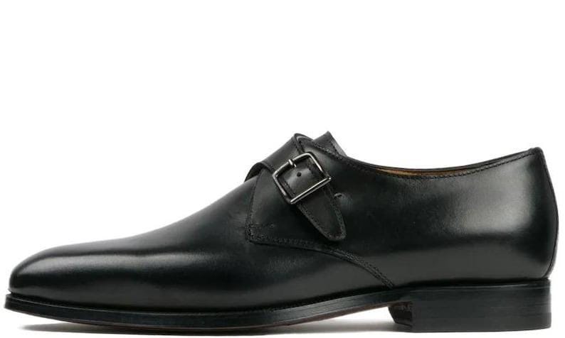May include: A single black leather monk strap shoe with a silver buckle. The shoe has a pointed toe and a low heel. The leather appears smooth and polished, suggesting a formal or dressy style. The shoe is designed for a right foot.