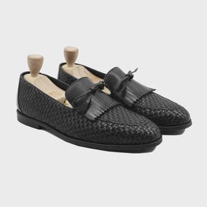 Peut inclure: Une paire de mocassins en cuir noir avec une texture tressée sur le dessus, un détail de franges kiltie et un nœud décoratif. Chaque chaussure est maintenue par un embauchoir en bois clair inséré à l'intérieur pour conserver sa forme.