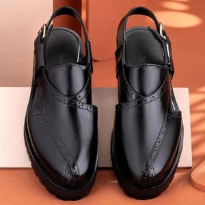 May include: A pair of black leather sandals featuring a closed-toe design with decorative brogue-style perforations along the vamp. The sandals include an adjustable ankle strap with a gold-tone metal buckle and a thick, textured black rubber sole.