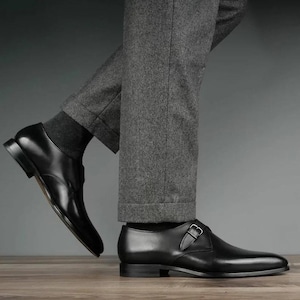 May include: Black leather dress shoes, one Oxford and one monk strap with a silver buckle. The shoes are worn with grey trousers and dark grey socks. The shoes are on a wooden surface.