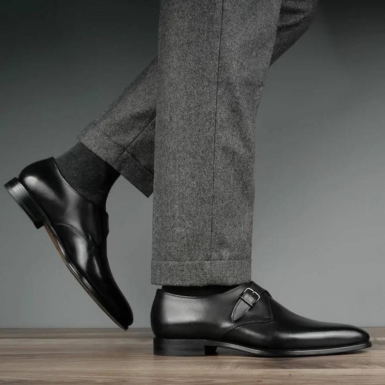 May include: Black leather dress shoes, one Oxford and one monk strap with a silver buckle. The shoes are worn with grey trousers and dark grey socks. The shoes are on a wooden surface.
