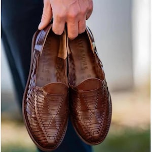 May include: A pair of brown leather huarache shoes featuring a woven upper design with a closed toe and an open heel area. The interior of the shoes displays the brand name OLYA & ACHILE printed on the footbed along with the size number 43.