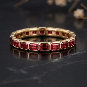 May include: A gold eternity band featuring a continuous row of rectangular, step-cut red rubies set in a bezel setting. The ring displays a polished metallic finish and a uniform arrangement of vibrant red gemstones encircling the entire band.