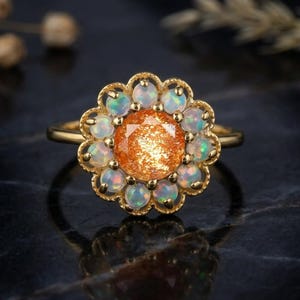 May include: A gold ring featuring a central round faceted orange sunstone surrounded by a halo of twelve small round white opals. The setting has a scalloped edge design, and the band is a simple gold wire, creating a floral-inspired appearance.