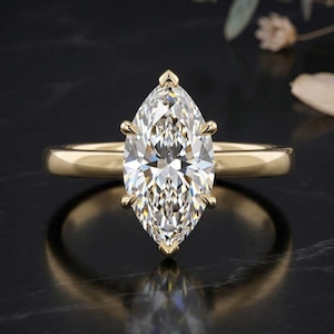 May include: A yellow gold ring featuring a large, marquise-cut clear gemstone set in a six-prong setting. The band is smooth and polished, showcasing a classic design with a pointed oval stone that reflects light across its faceted surface.