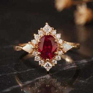 May include: A gold ring featuring a central oval-cut deep red ruby surrounded by a halo of small round white diamonds. Two triangular white opals are set on the sides of the band, with additional small square-cut diamonds at the top and bottom.