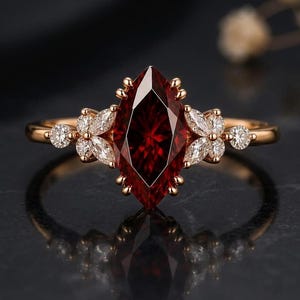 May include: A rose gold ring featuring a central marquise-cut deep red gemstone. The setting is accented by smaller marquise-cut and round-cut clear white stones arranged in a floral-like cluster on both sides of the main red stone.