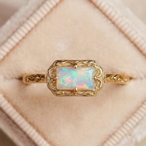 May include: A gold-colored ring with an opal stone. The rectangular opal is set in a decorative, filigree-style frame. The ring band features an ornate design. The ring is displayed in a soft, pink ring box.