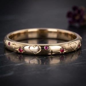 May include: A gold band ring featuring engraved heart motifs and star-shaped settings, each containing a small, round, vibrant red gemstone. The polished metal surface reflects light, highlighting the alternating pattern of hearts and stars.