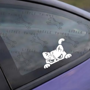 Pode incluir: Autocolante de carro em vinil branco com um gato de desenho animado com uma expressão zangada. O autocolante está numa janela de carro com um padrão de ilustrações de cavalos a correr e as palavras "LITTLE THINGS".