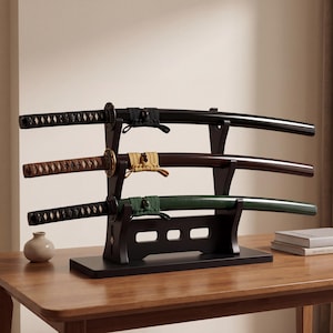 May include: Three Japanese katana swords displayed on a black wooden stand. The swords have black, brown, and green sheaths, with decorative handles and fittings. The stand is on a wooden table.