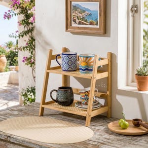 Puede incluir: Una estanter&iacute;a de bamb&uacute; de dos niveles exhibe cuatro tazas de cer&aacute;mica con dise&ntilde;os coloridos. La estanter&iacute;a superior contiene una taza azul estampada y una taza con un dise&ntilde;o de sol y olas. La estanter&iacute;a inferior tiene una taza moteada oscura y una taza estampada con un platillo.