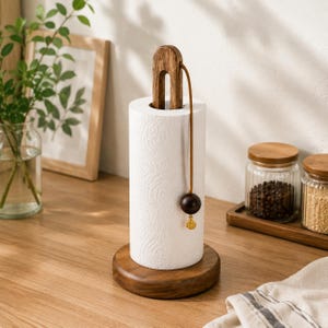Puede incluir: Un portarrollos de papel de cocina de madera con acabado marrón oscuro. El soporte tiene un asa en forma de U, un cordón de cuero y una bola decorativa de madera con un dije dorado. El rollo de papel de cocina blanco se asienta sobre una base redonda de madera.
